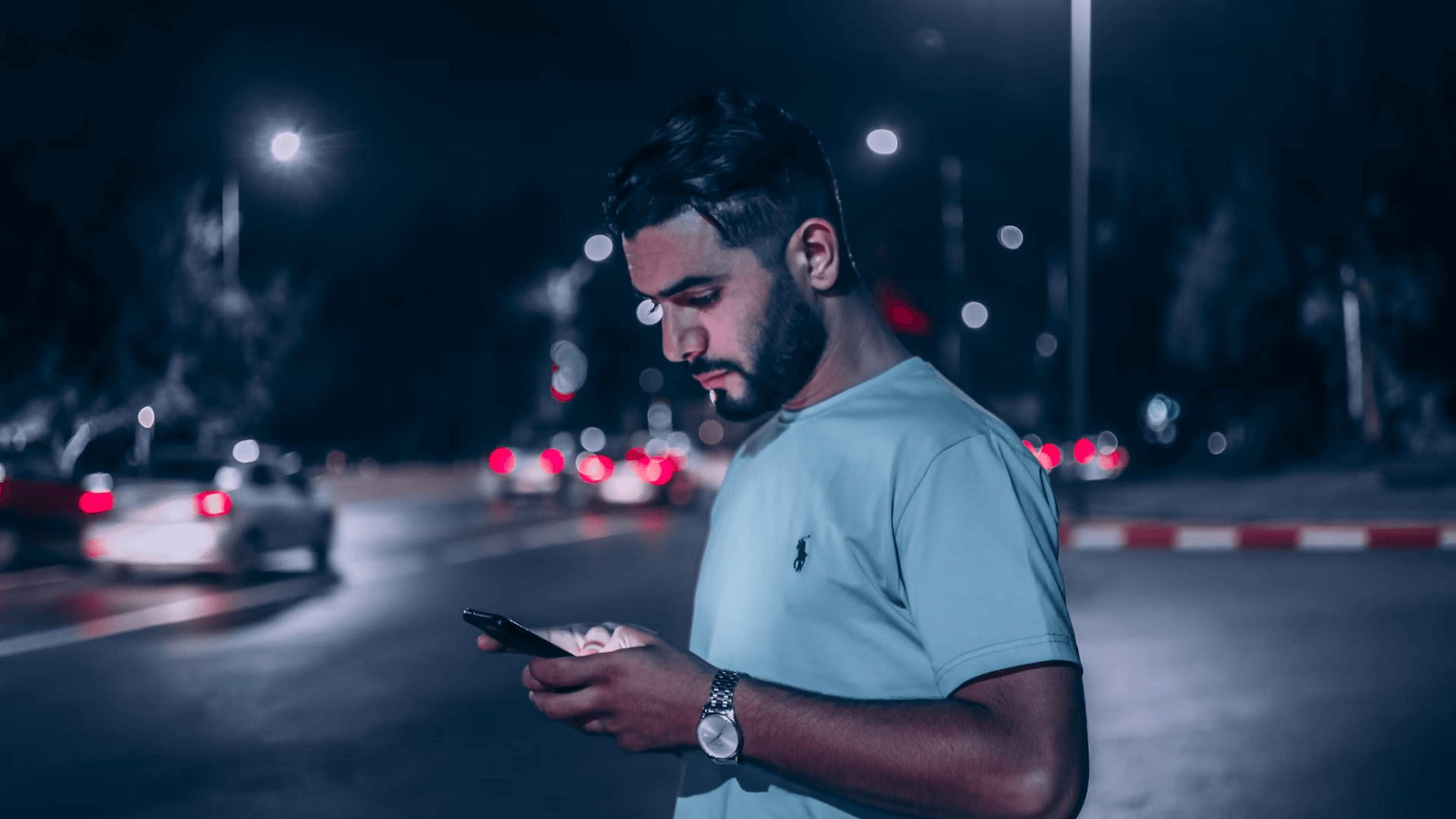 A young man standing in a parking lot at night looking at his phone
