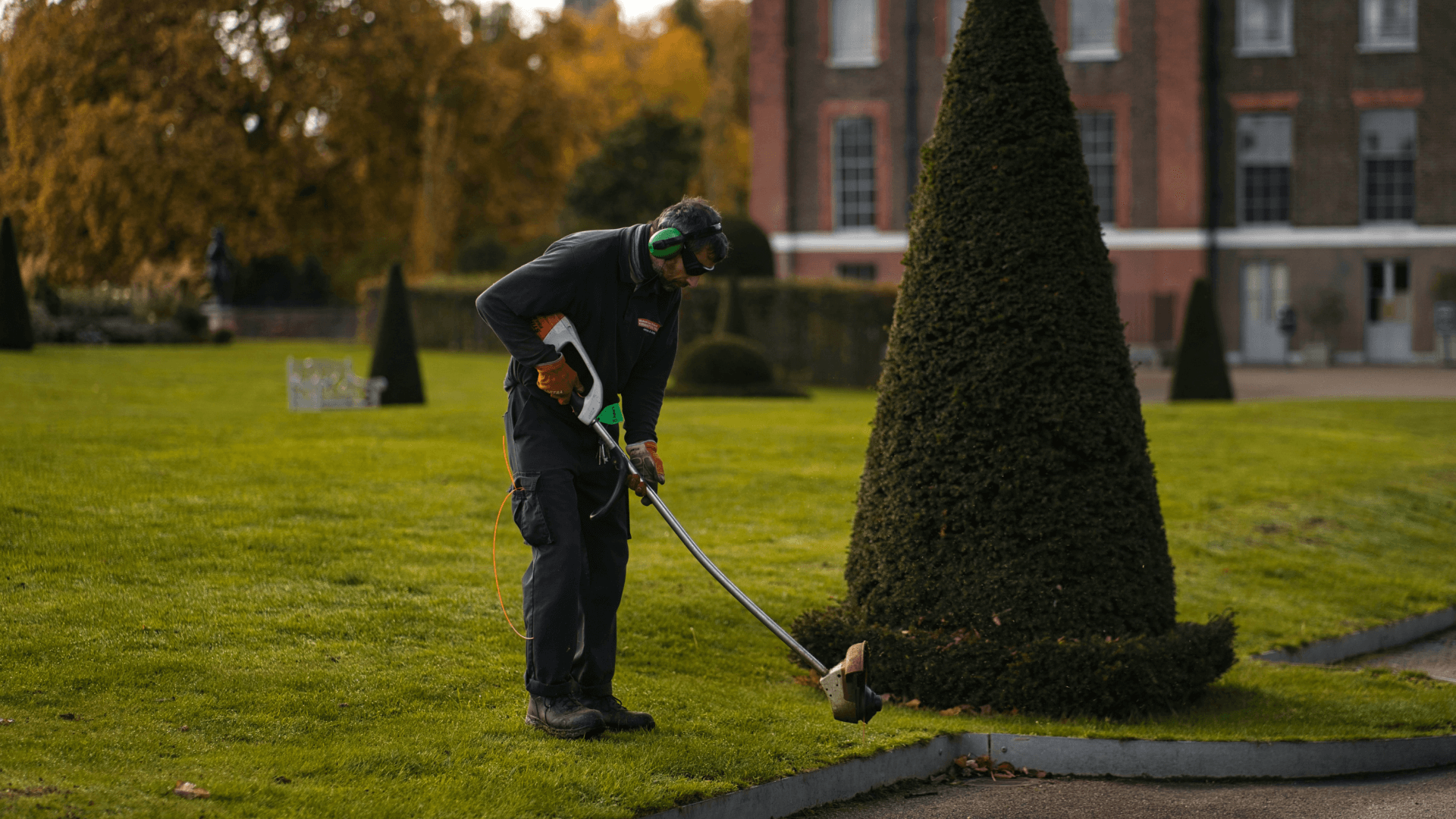 A man in a navy jumpsuit using a weed eater/lawn trimmer on a lawn