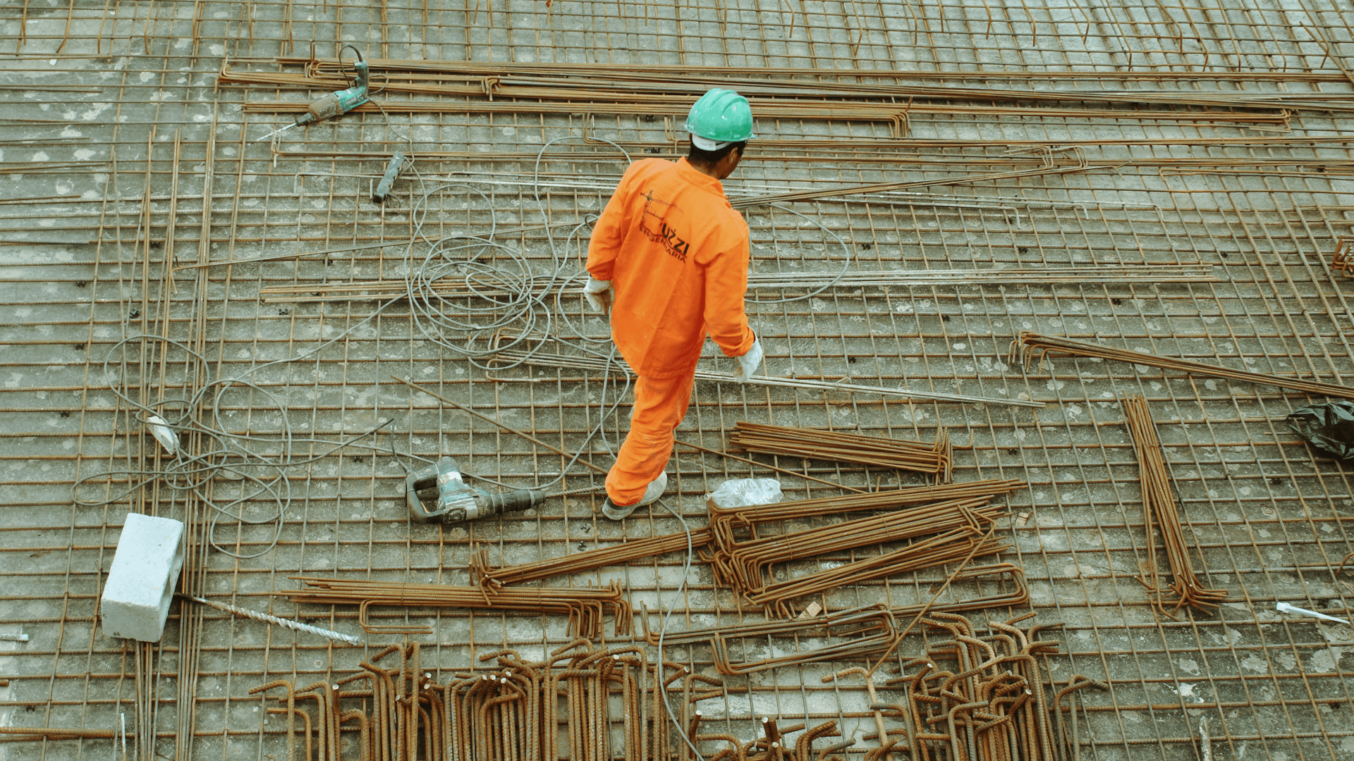 obrero de la construcción vestido con un mono naranja brillante y un casco verde, caminando por una cimentación de rejilla metálica, que está cubierta de barras de refuerzo y herramientas de construcción.