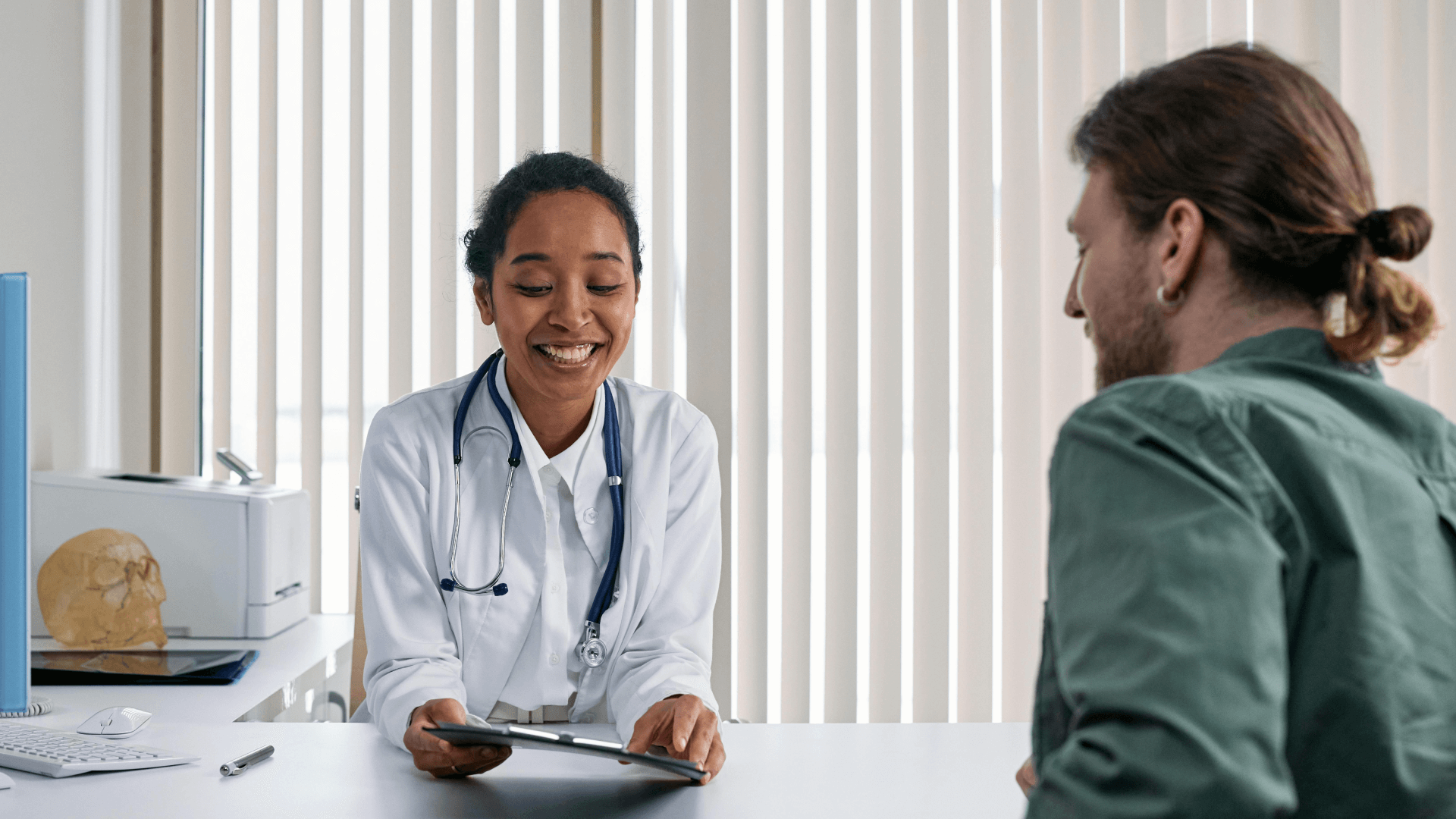 Un amable médico, con bata blanca y estetoscopio, sonríe sentado frente a un paciente en un despacho. El médico sostiene una tableta, probablemente revisando o compartiendo información médica con el paciente, que está sentado de espaldas a la cámara. El paciente lleva el pelo largo recogido en un moño y viste una camisa verde. El ambiente es de colaboración y tranquilidad.