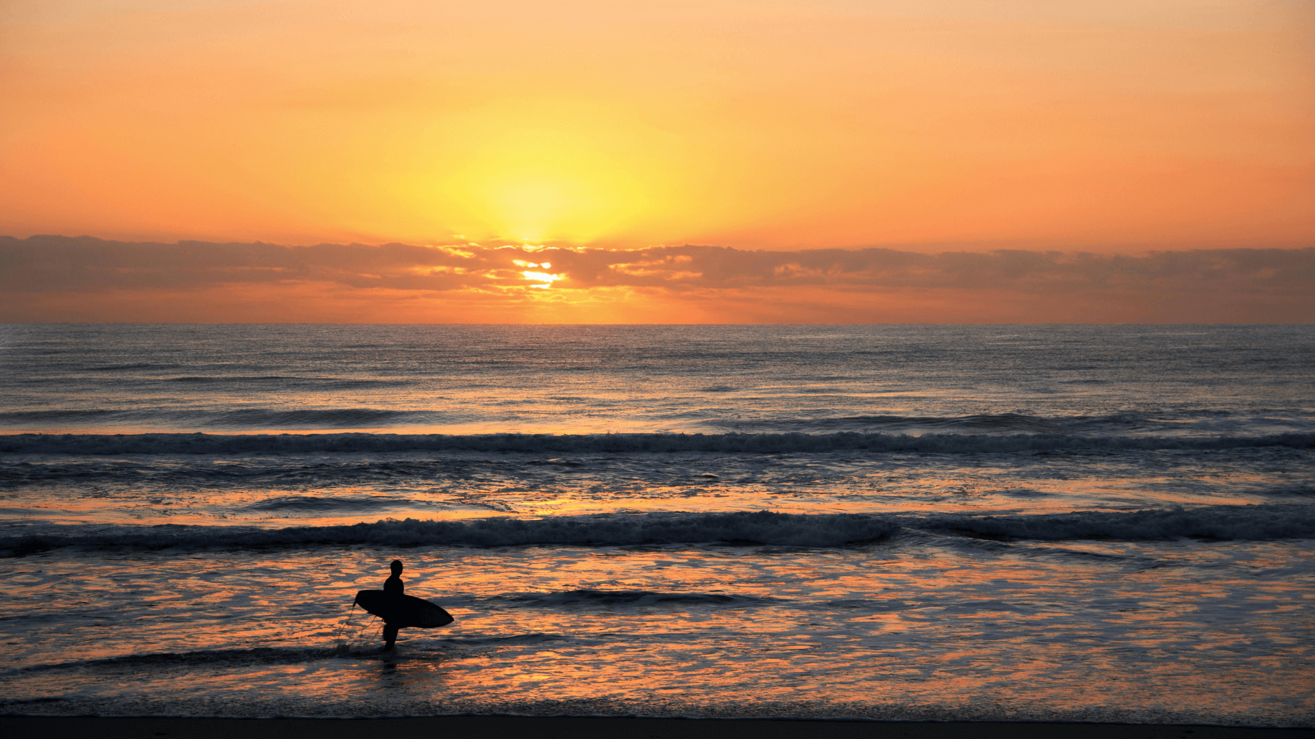 Una foto de una cálida puesta de sol sobre el océano. En la esquina inferior izquierda hay un surfista con una tabla mirando al horizonte.