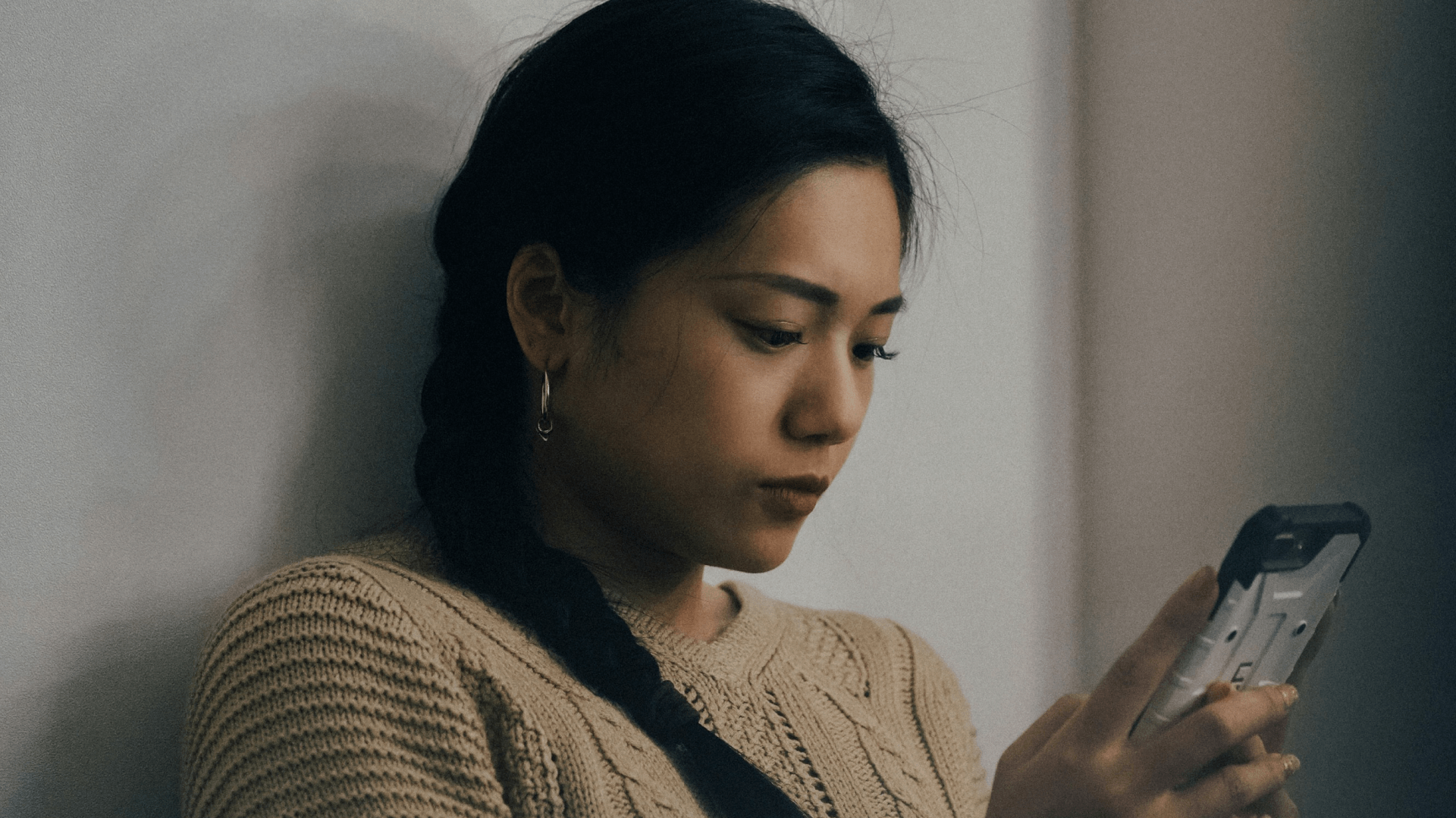 A photo of a young woman with dark hair braided over her shoulder, intently looking at her phone. She is wearing a beige knit sweater and simple hoop earrings. Her expression is calm and focused as she interacts with her device, she is seated against a neutral-colored wall. The soft lighting creates a thoughtful, reflective mood.