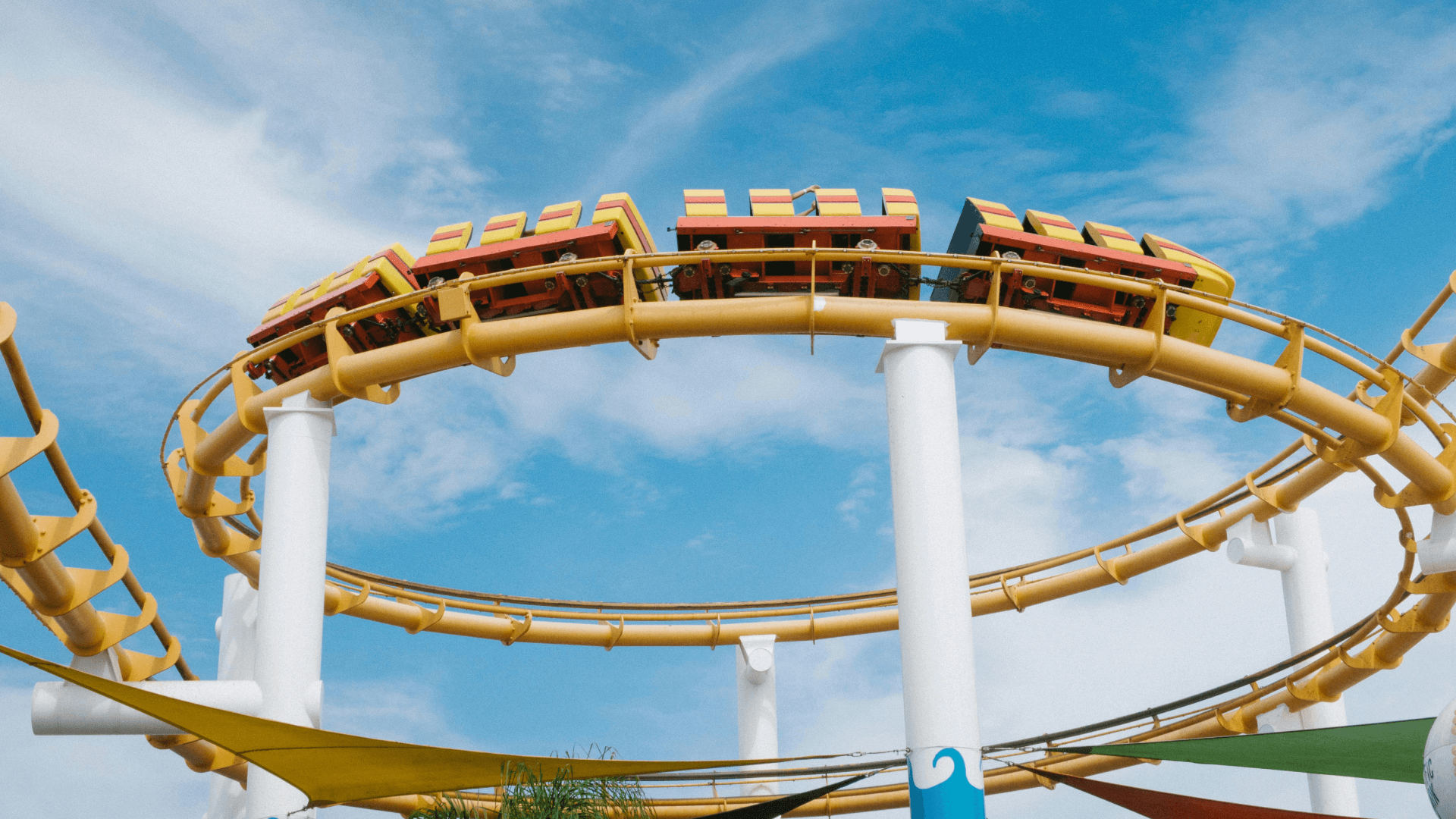 A brightly colored roller coaster, with red, yellow, and orange cars, traveling along a curving yellow track. The track is supported by tall white columns, and the ride takes place under a clear blue sky with light clouds. There are colorful canopies visible at the bottom of the image, possibly part of a theme park or fairground setting. The perspective looks up toward the roller coaster, capturing the motion and excitement of the ride.