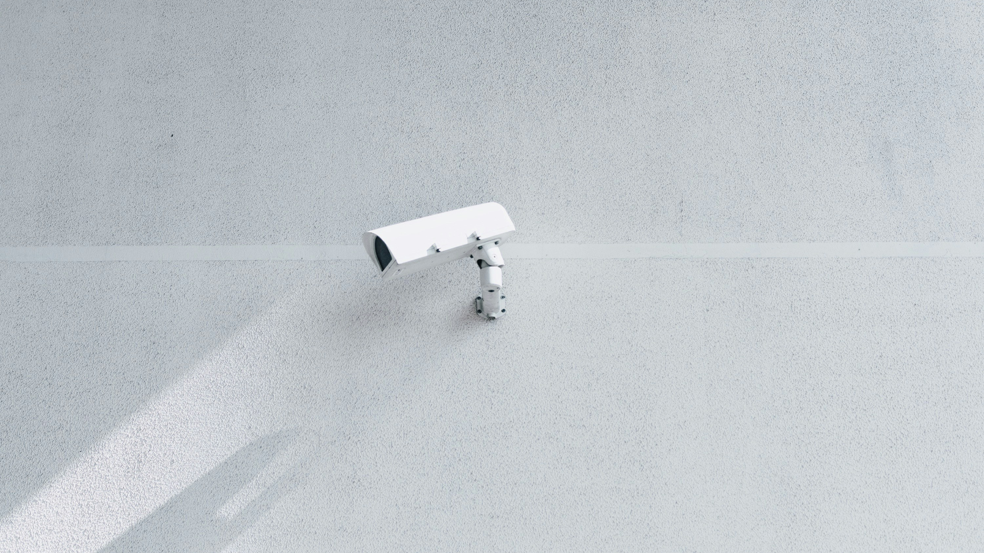 A white surveillance camera mounted on a plain white wall, capturing its surroundings.