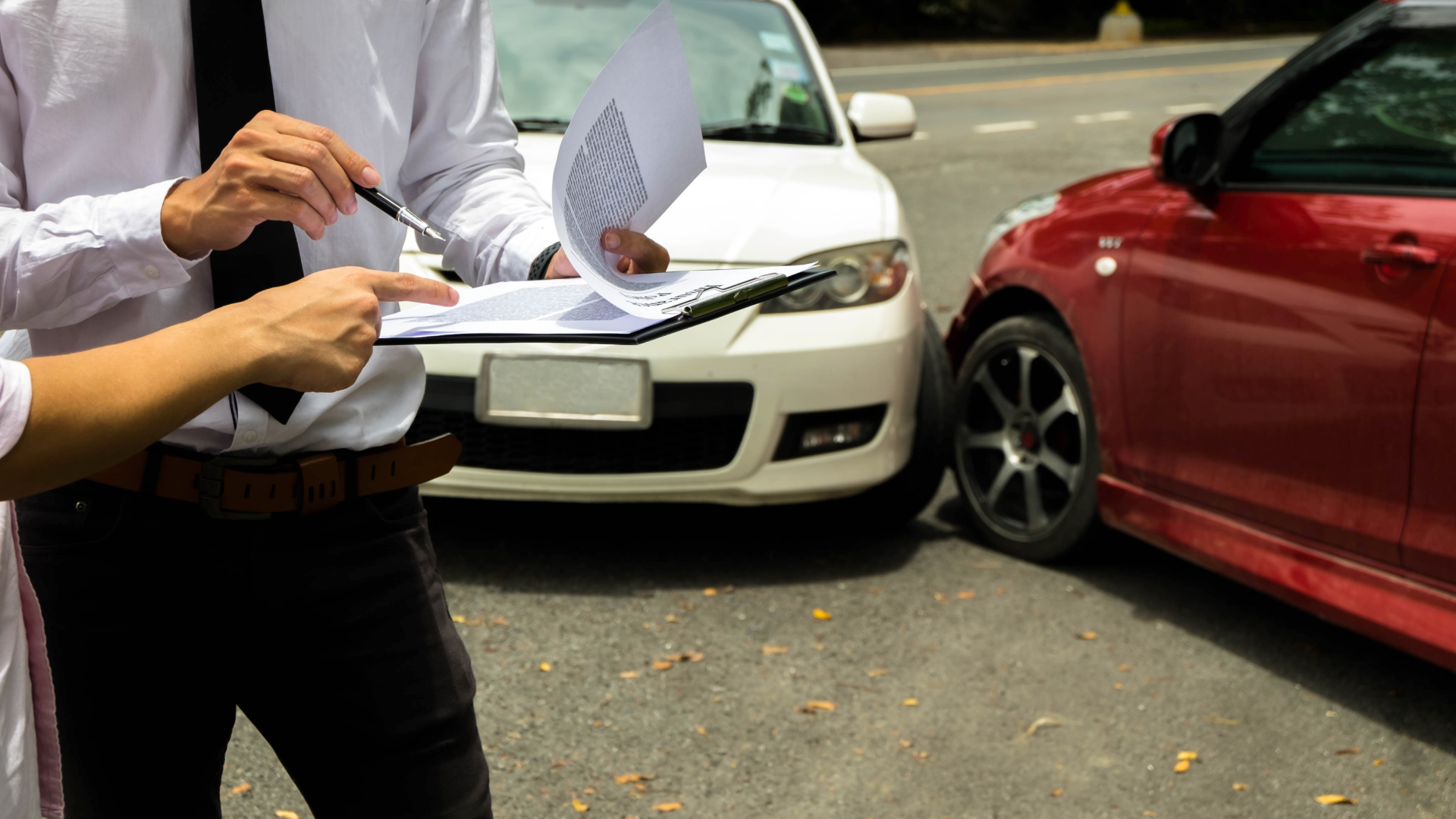 An insurance adjuster discussing paperwork with a client at the scene of a car accident, with two cars involved in a collision in the background.