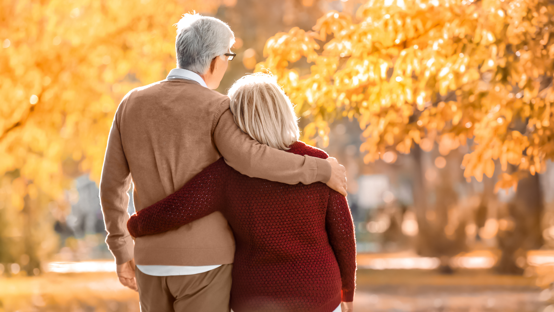 An elderly couple walking together in a park during autumn, with the man’s arm around the woman’s shoulder, showing affection and support.