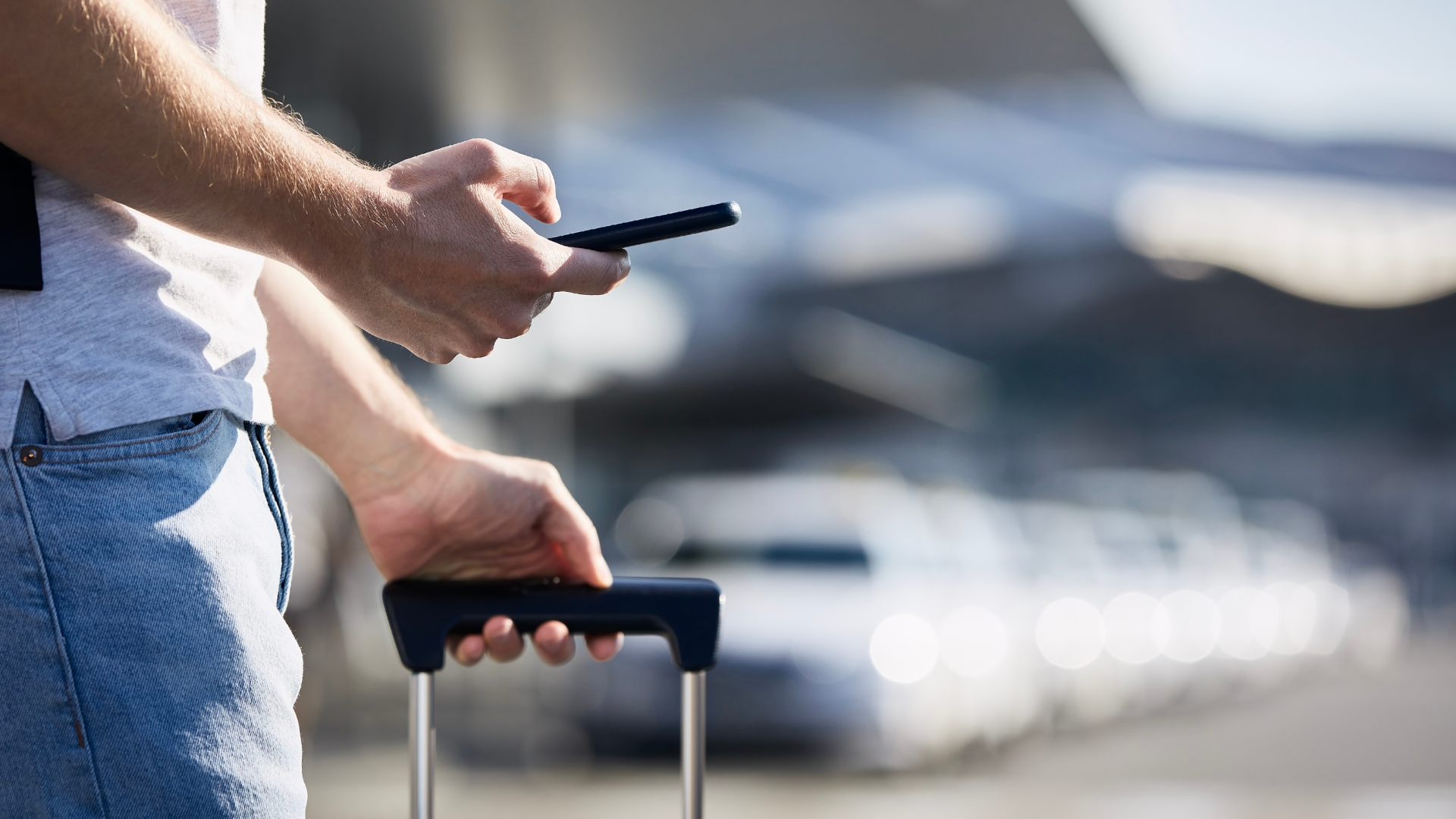 A man holding a suitcase and looking at his phone while waiting for a rideshare in an airport parking area.