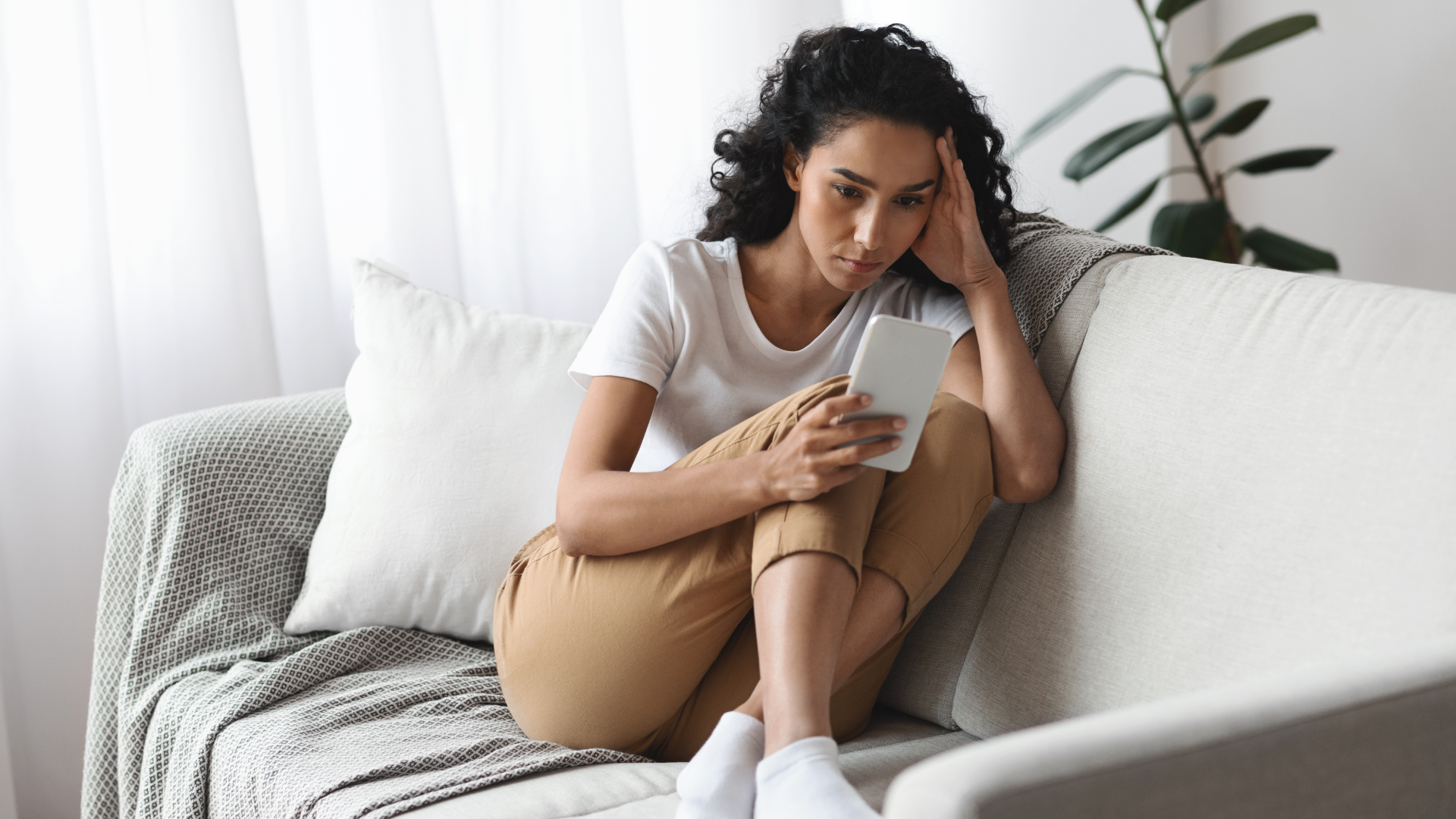 A young woman with curly hair sitting on a couch, looking at her phone with a concerned expression, resting her head on her hand in a moment of distress.