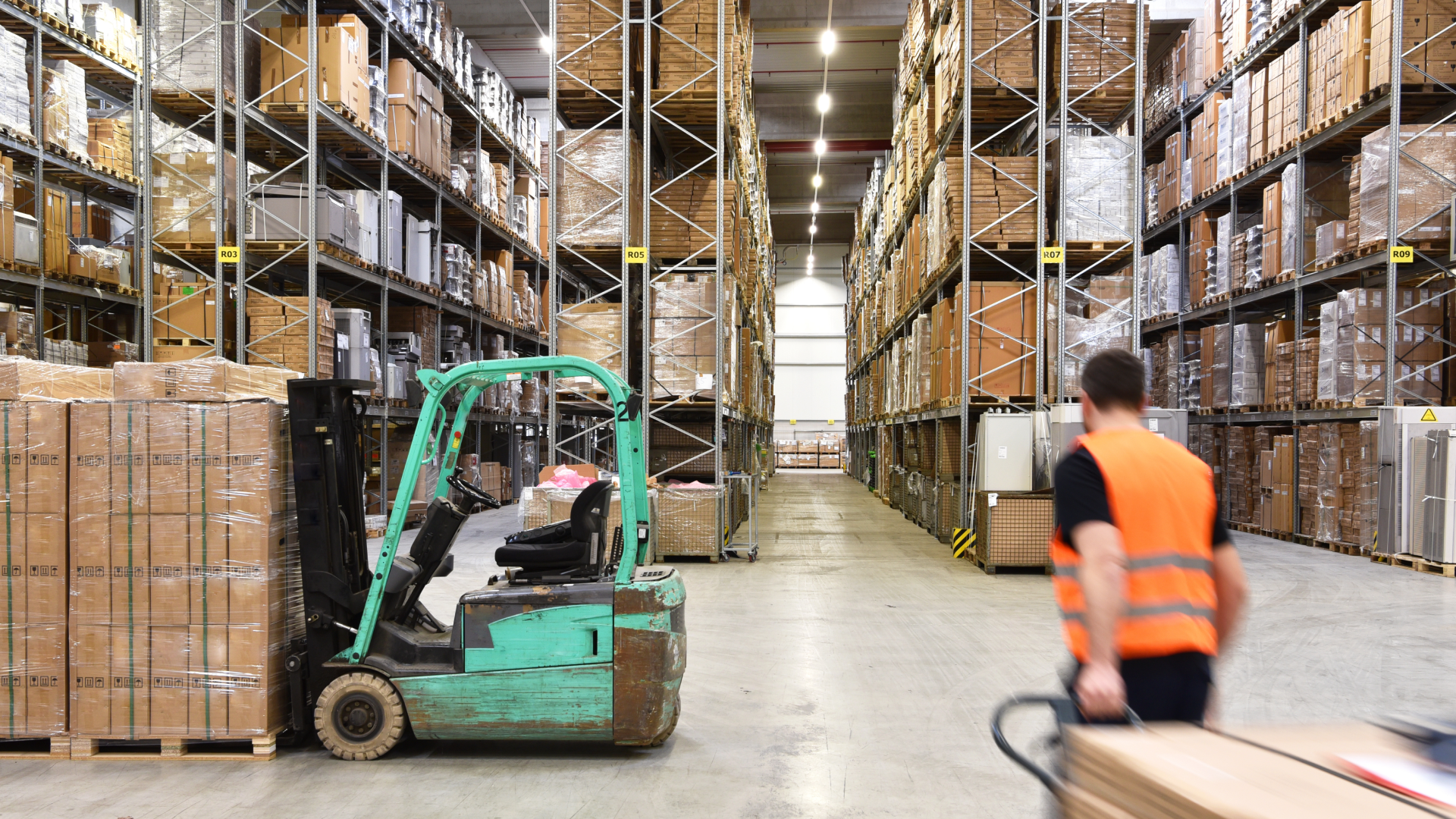 Warehouse worker in safety vest walking past forklift and stacked pallets in a high-shelving storage facility