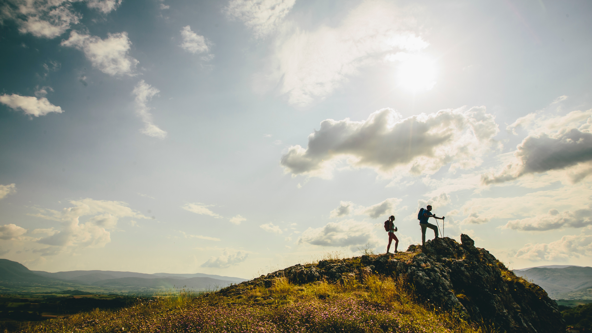 Two hikers with backpacks ascend a rocky hilltop under a bright sun, silhouetted against a scenic mountain landscape.