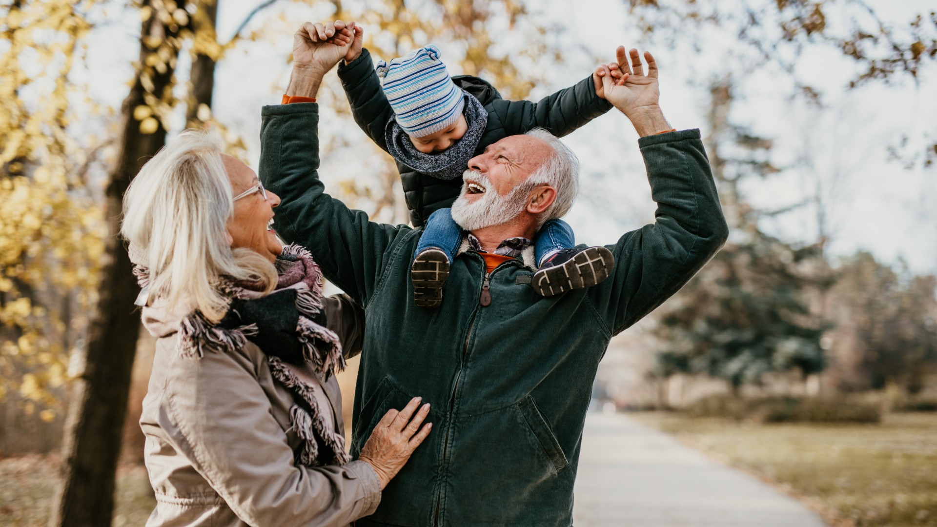 Happy grandparents playing with their grandchild outdoors, with the grandfather lifting the child on his shoulders while the grandmother smiles beside them on a fall day.