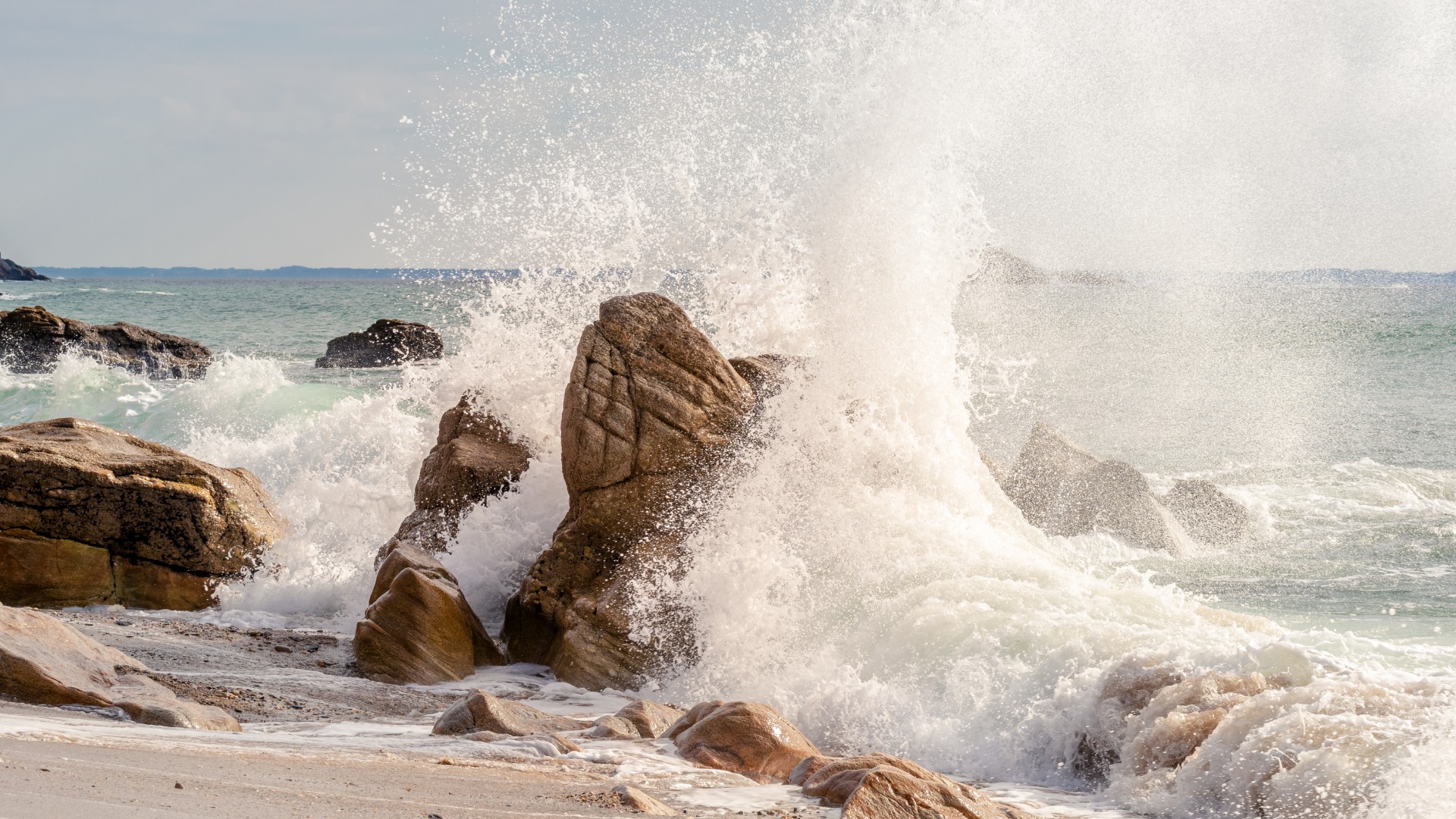 Ocean waves crashing against large rocks on a sandy shore.