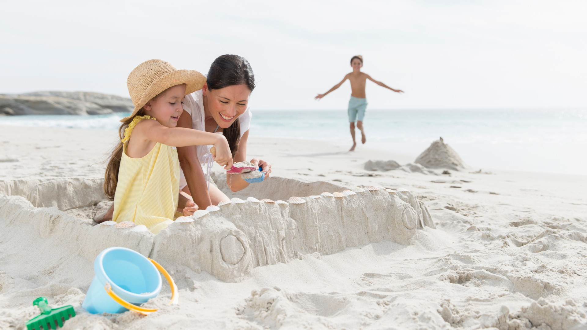 Una madre y su hija pequeña construyen un castillo de arena en una playa soleada mientras un niño corre hacia ellas cerca de la orilla.