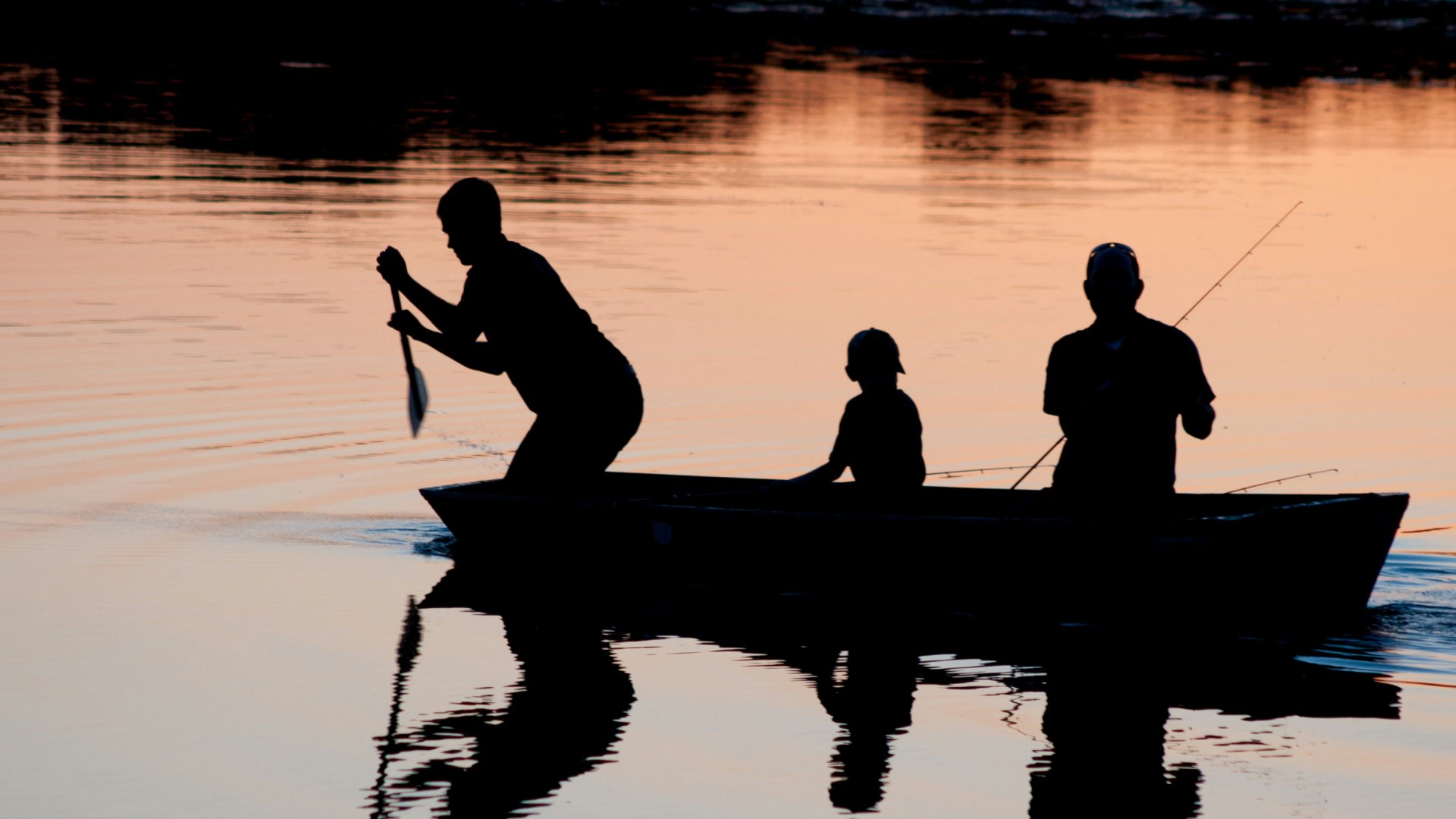 Silhouettes of three people in a small boat fishing on calm water at sunset.