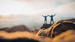 Two people standing on a rocky mountain overlook with arms raised.