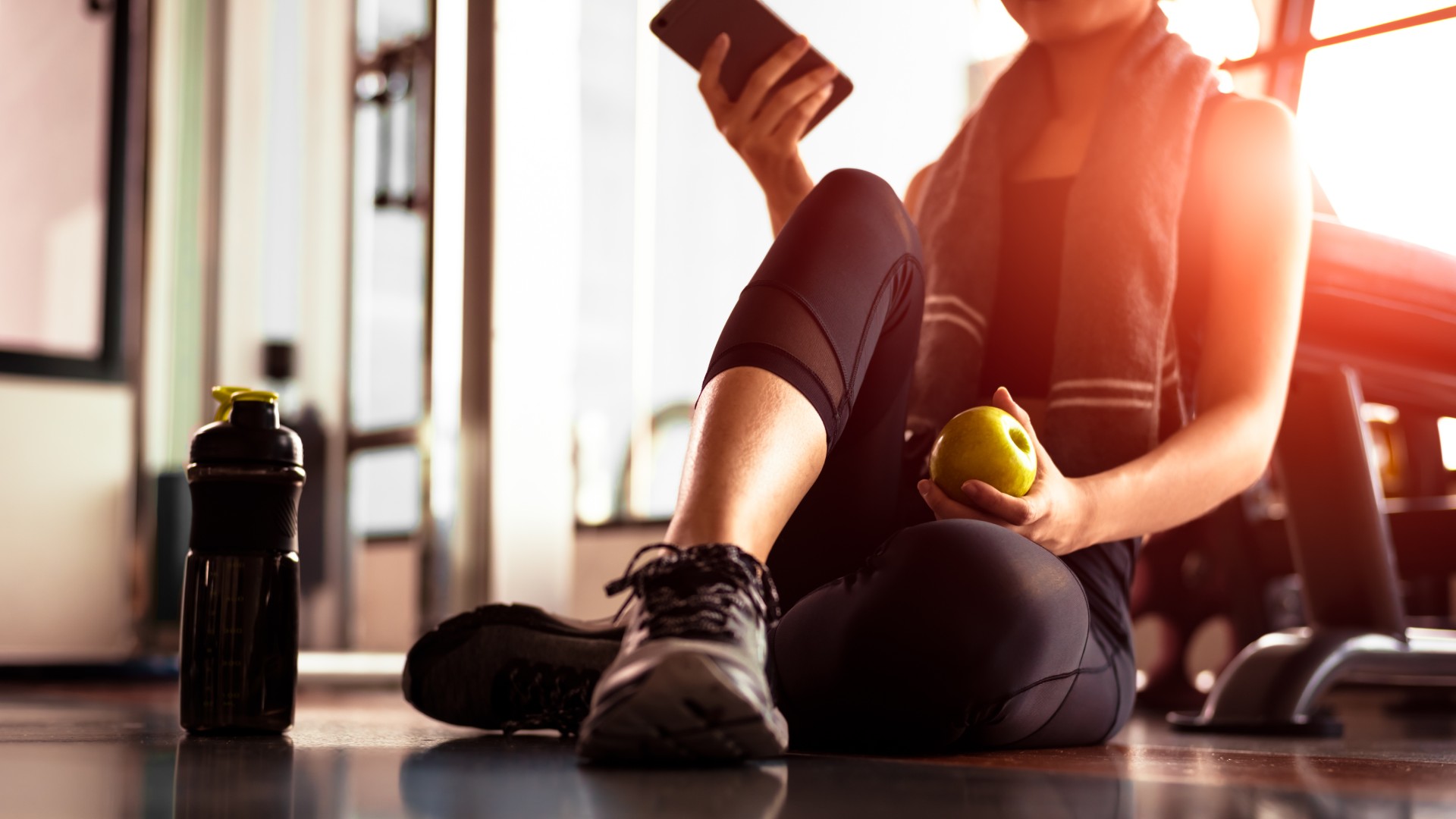 Person sitting on a gym floor holding a phone and an apple next to a water bottle.