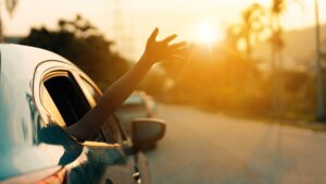 Person’s arm extended out of a car window toward the sun while driving down a road at sunset.