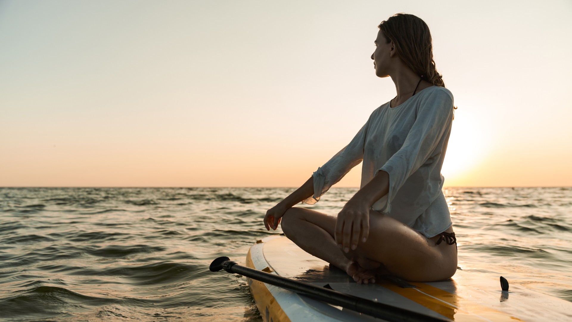 Woman sitting cross-legged on a paddleboard in the ocean at sunset, looking out over the water.