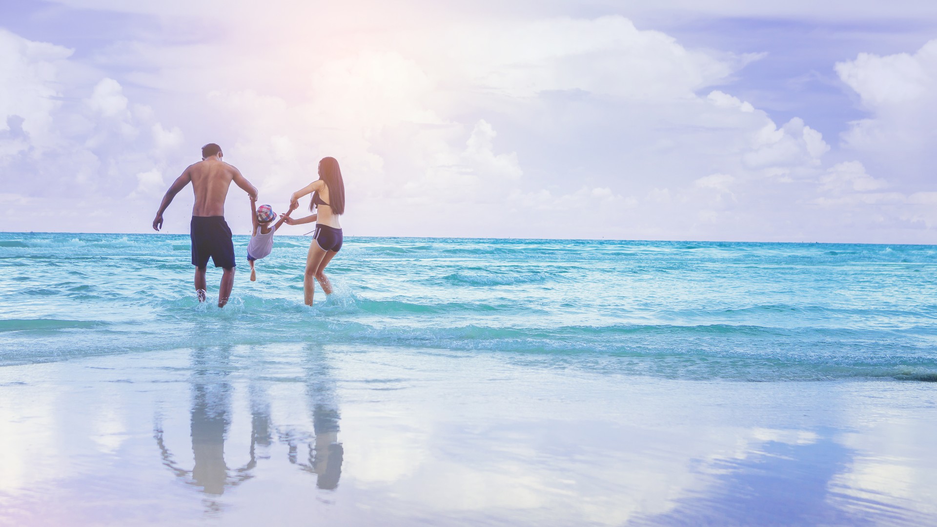 Parents holding their child’s hands while playing together in shallow ocean water at the beach.