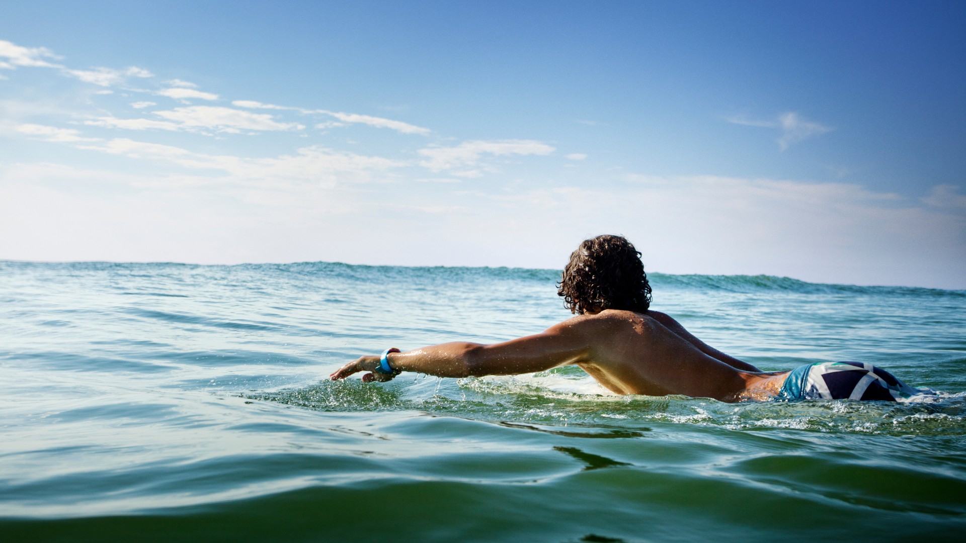 Person paddling on a surfboard in calm ocean water under a bright blue sky.