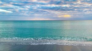 Calm turquoise ocean water meeting a sandy beach beneath a cloudy sunrise sky.