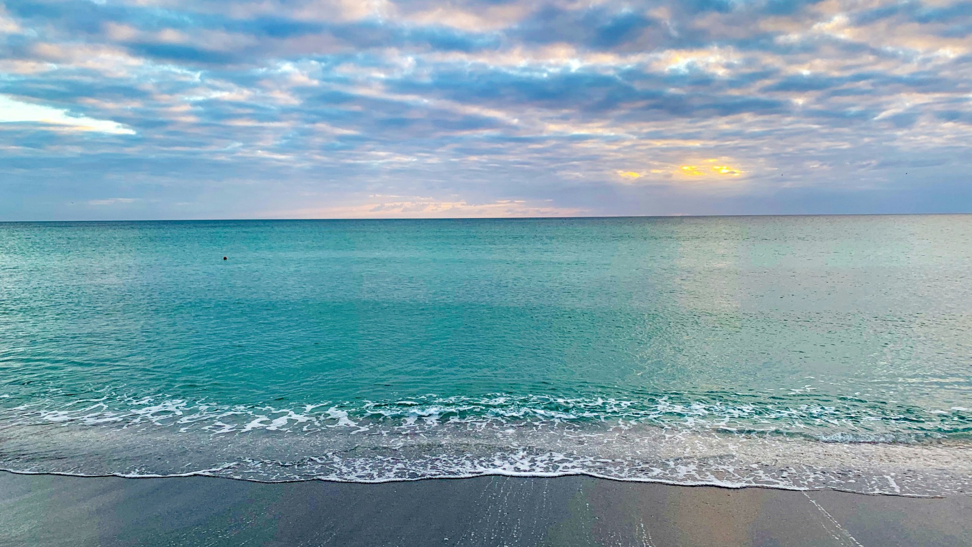 Calm turquoise ocean water meeting a sandy beach beneath a cloudy sunrise sky.