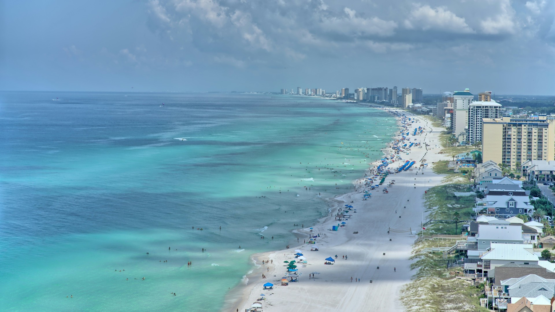 Aerial view of a crowded beachfront with turquoise ocean water, white sand, and high-rise buildings along the coast.