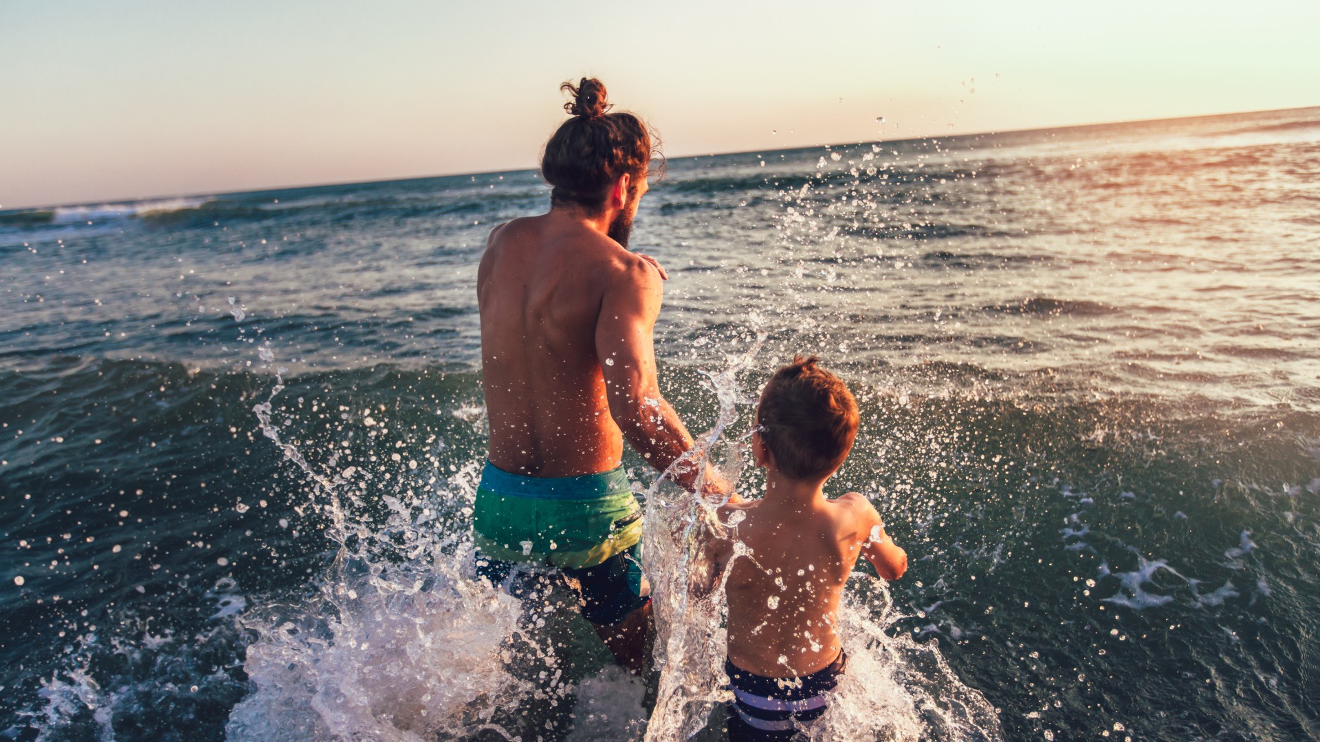 Father and son running into ocean waves together at sunset with water splashing around them.