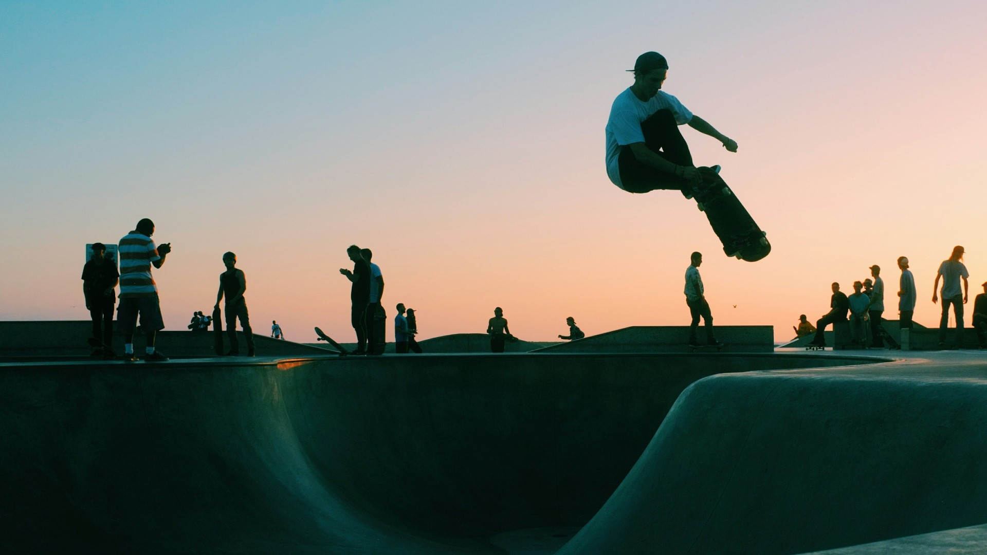 Skateboarder performing a high jump trick at a skatepark during sunset with other skaters in the background