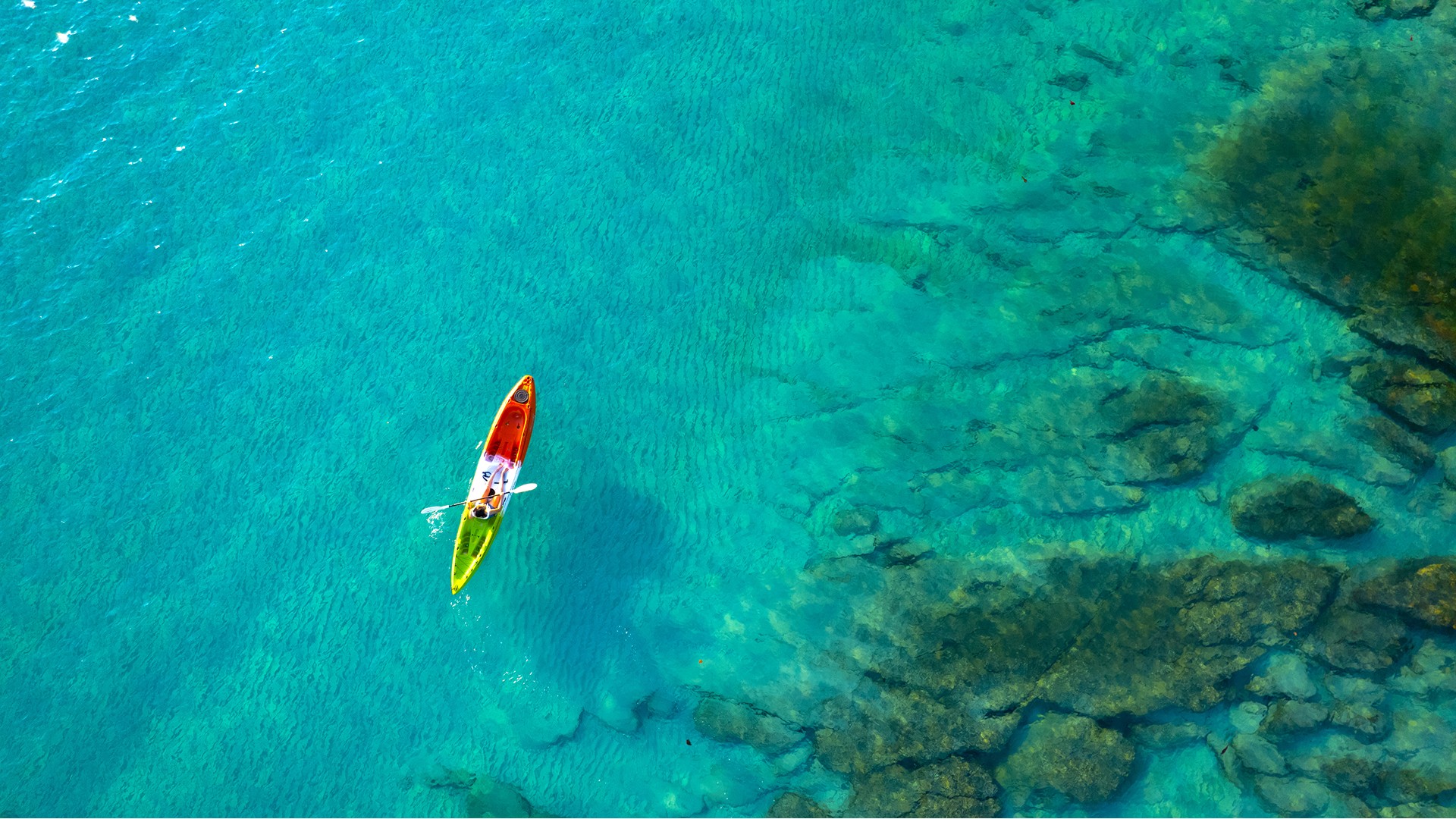Aerial view of a colorful kayak floating over clear turquoise water near a rocky shoreline.