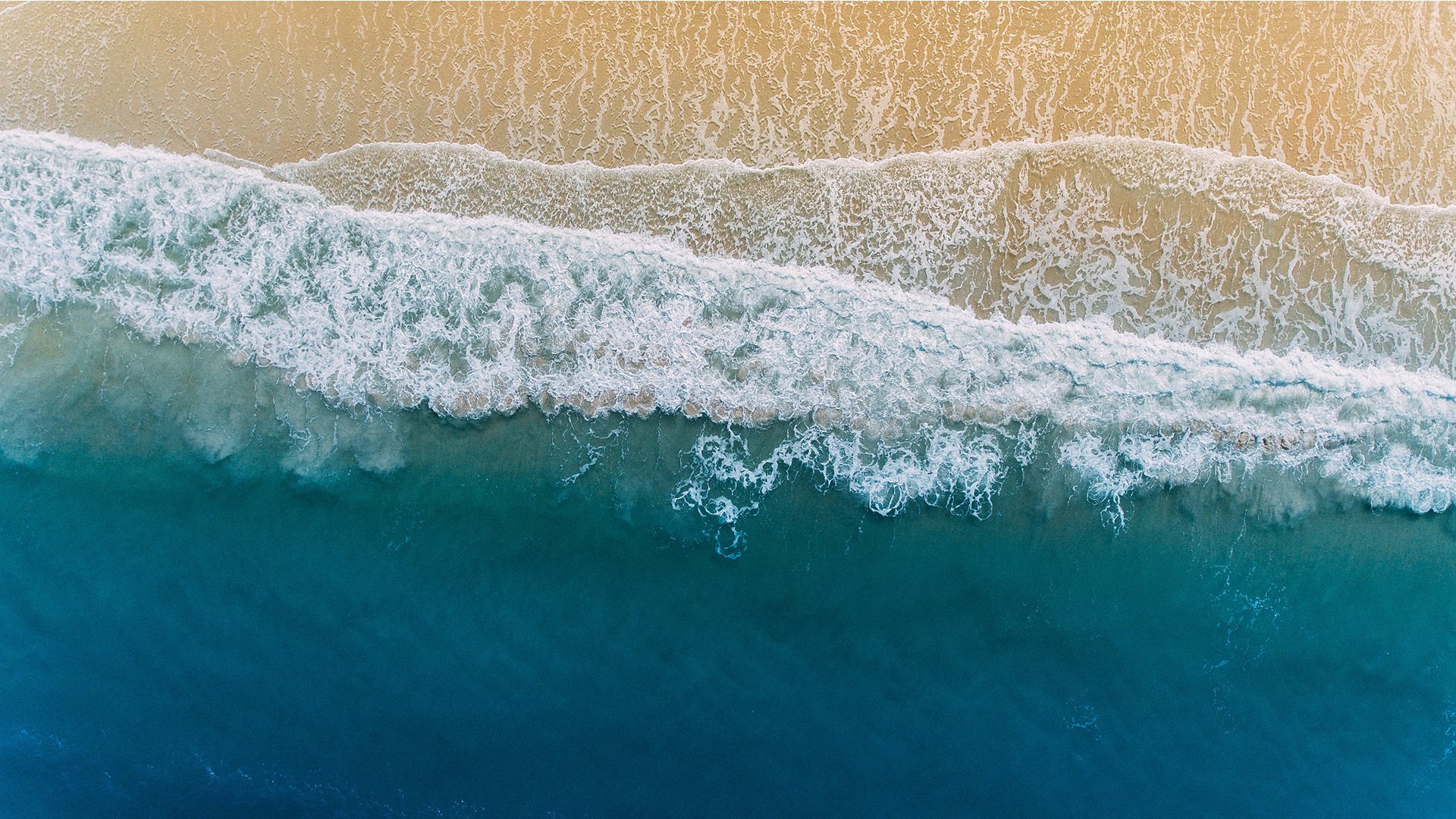 Aerial view of ocean waves rolling onto a sandy beach with turquoise water and white sea foam.