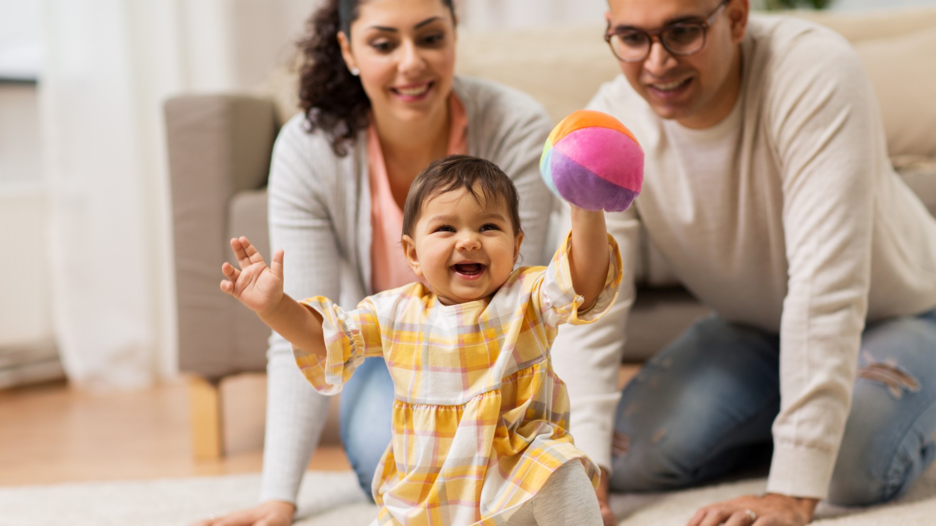 Smiling baby playing with a colorful ball while parents watch nearby in a living room.