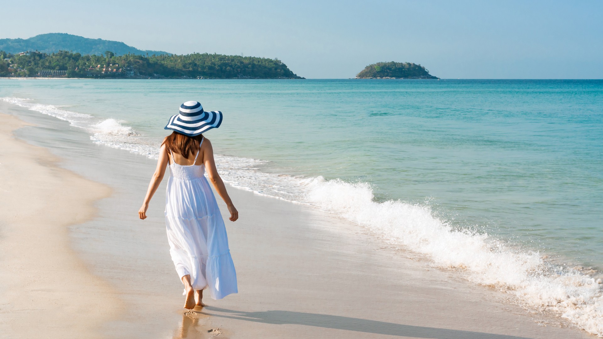 Woman in a white dress and wide-brim hat walking barefoot along a tropical beach shoreline.