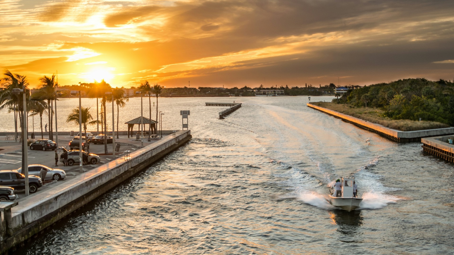 Boat speeding through a coastal inlet at sunset with palm trees, docks, and shoreline in the background.