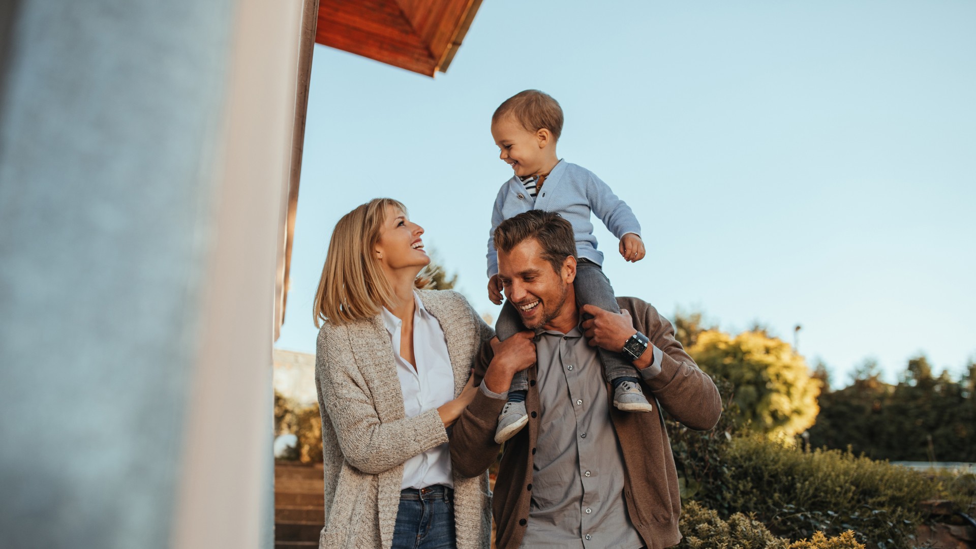 Happy parents walking outside with their young child sitting on the father’s shoulders, smiling together.