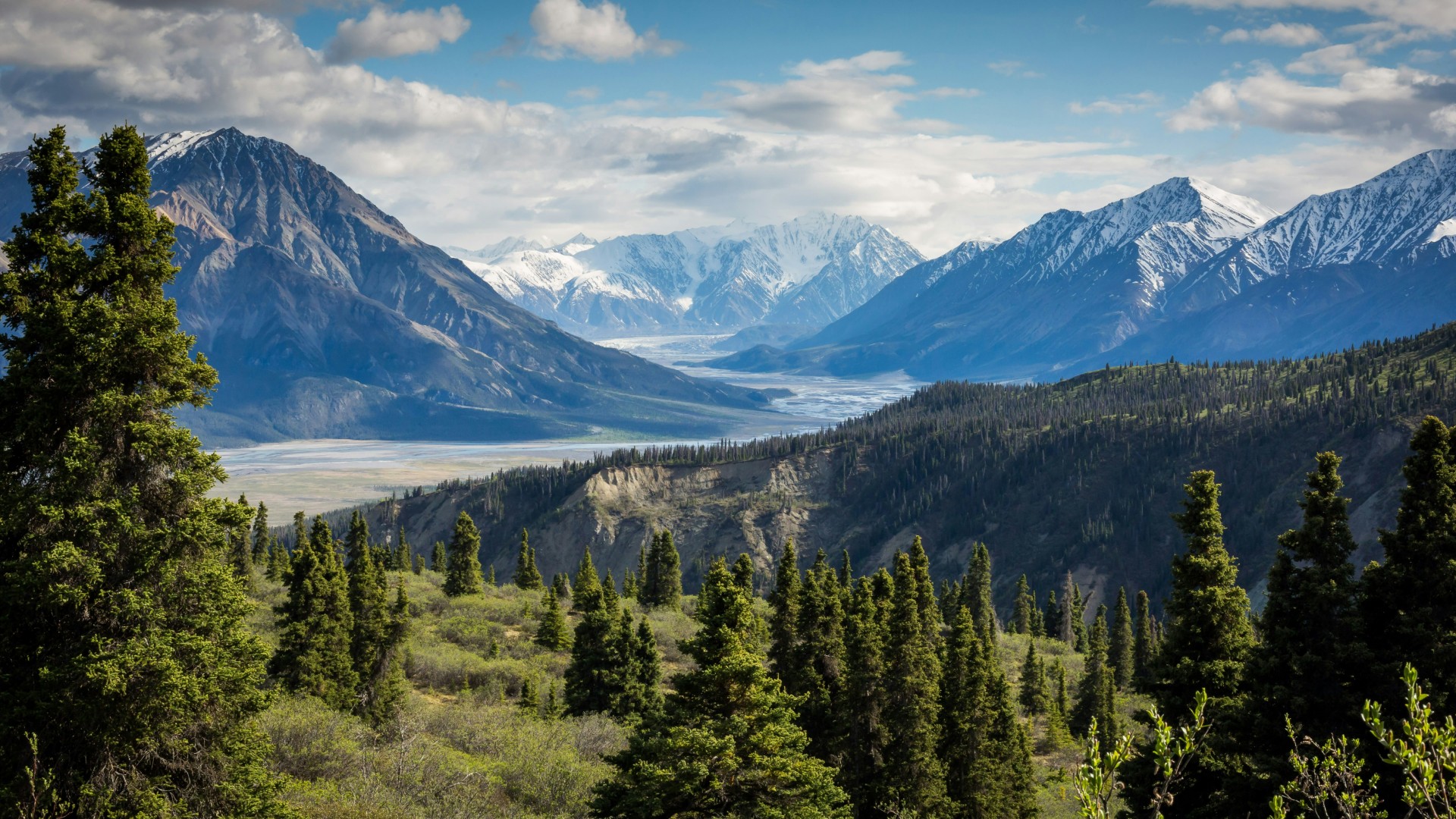 Scenic mountain valley with snow-capped peaks, evergreen forest, and a winding river under a partly cloudy sky.
