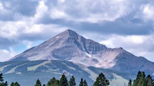 Snow-dusted mountain peak rising above evergreen forests under dramatic cloudy skies.