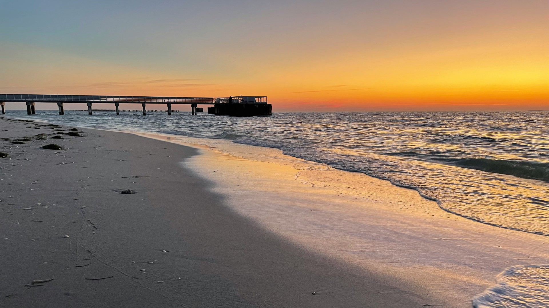 Sunset over a sandy beach with gentle waves and a long pier extending into the ocean.