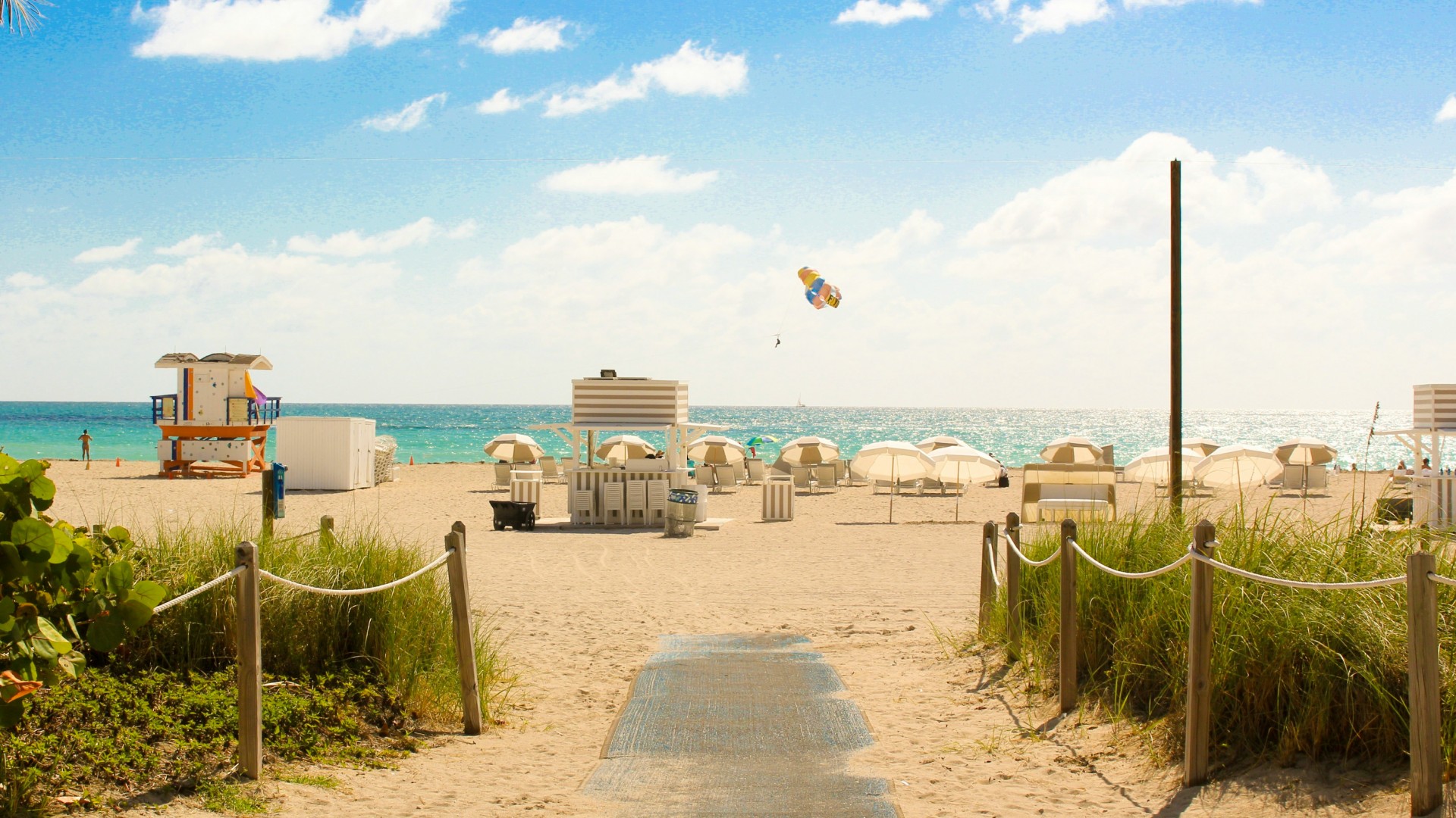 Sandy beach entrance leading to umbrellas, lifeguard towers, and turquoise ocean under a sunny sky.