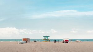 Wide sandy beach with a colorful lifeguard tower, a few beachgoers, and the ocean beneath a bright blue sky with soft clouds.