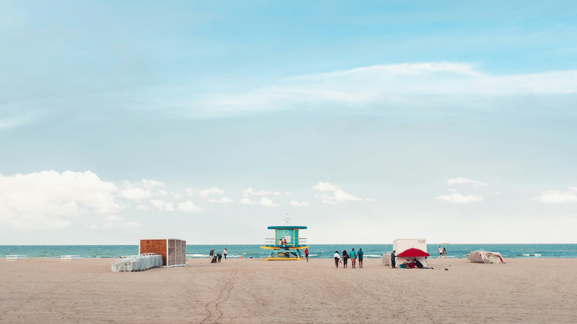 Wide sandy beach with a colorful lifeguard tower, a few beachgoers, and the ocean beneath a bright blue sky with soft clouds.
