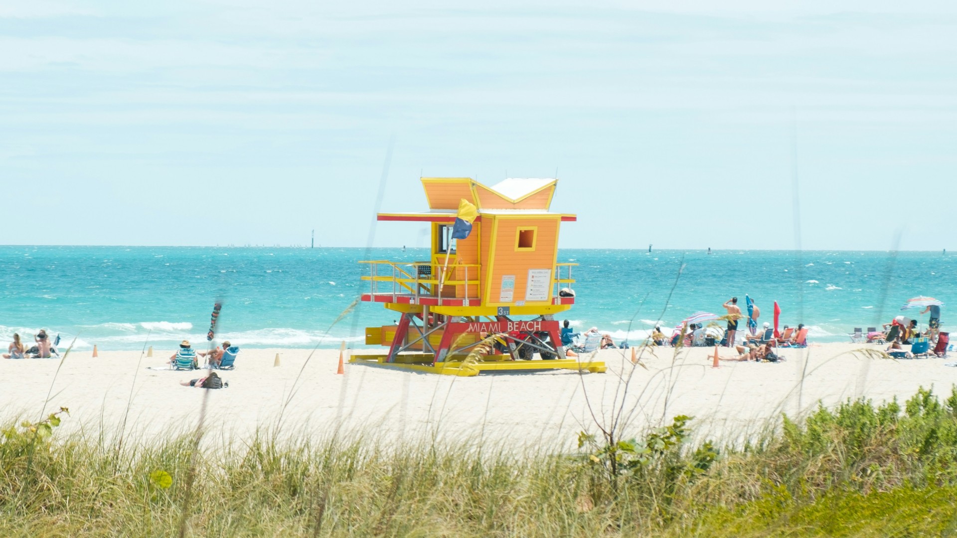 Colorful lifeguard tower on a sunny beach with beachgoers relaxing beside turquoise ocean waters.