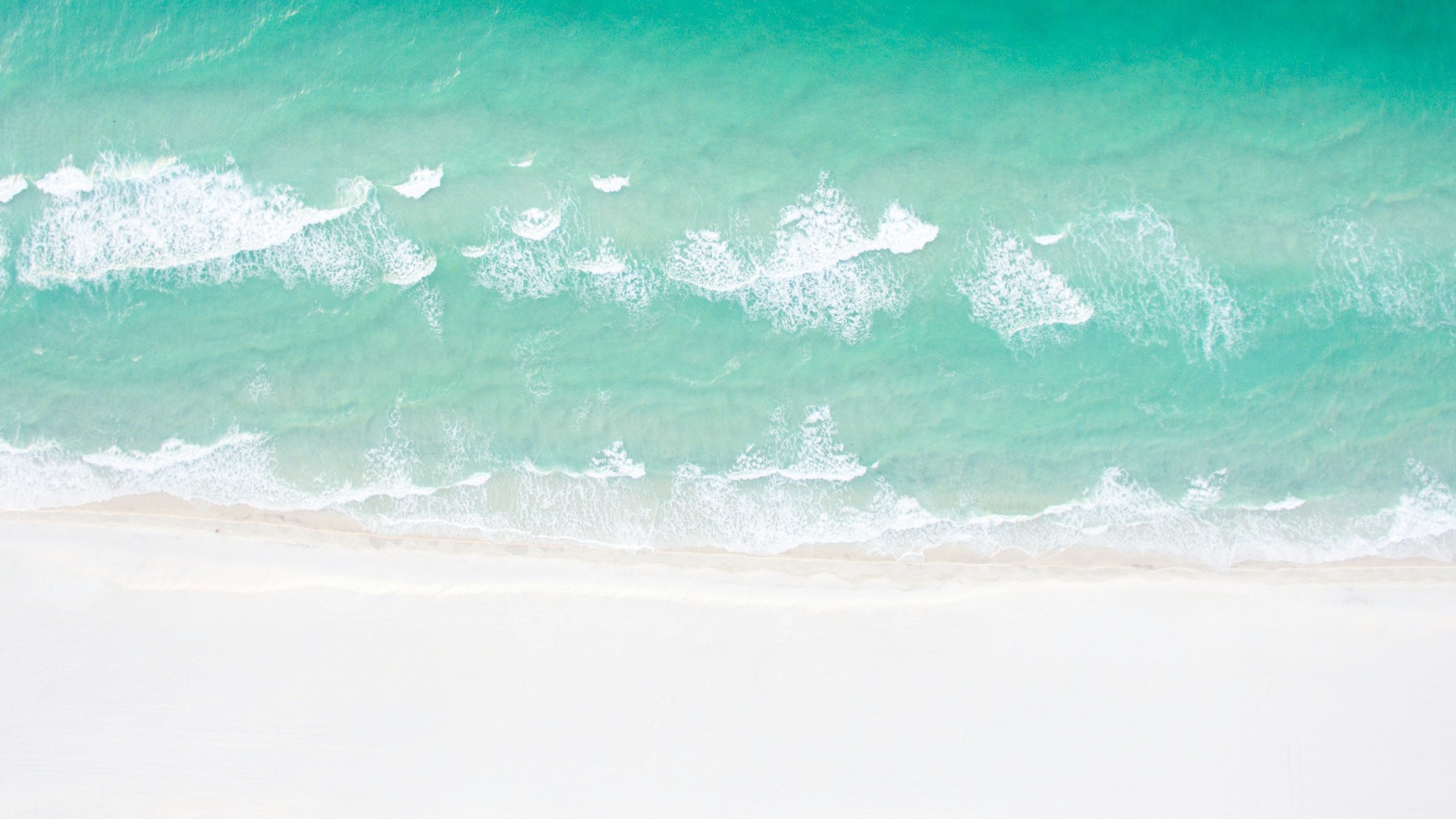 Aerial view of gentle turquoise ocean waves washing onto a wide white sand beach.