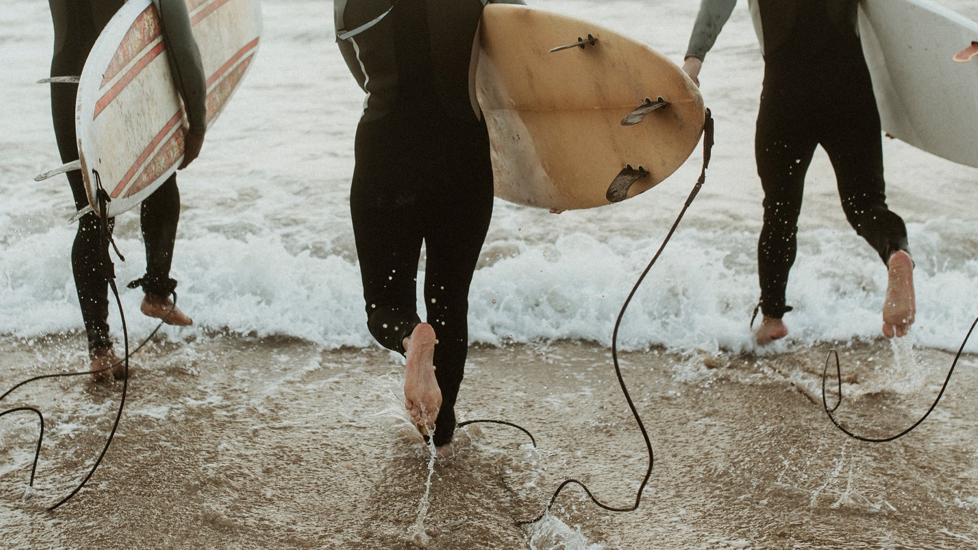 Surfers in wetsuits carrying surfboards as they run into the ocean through breaking waves.