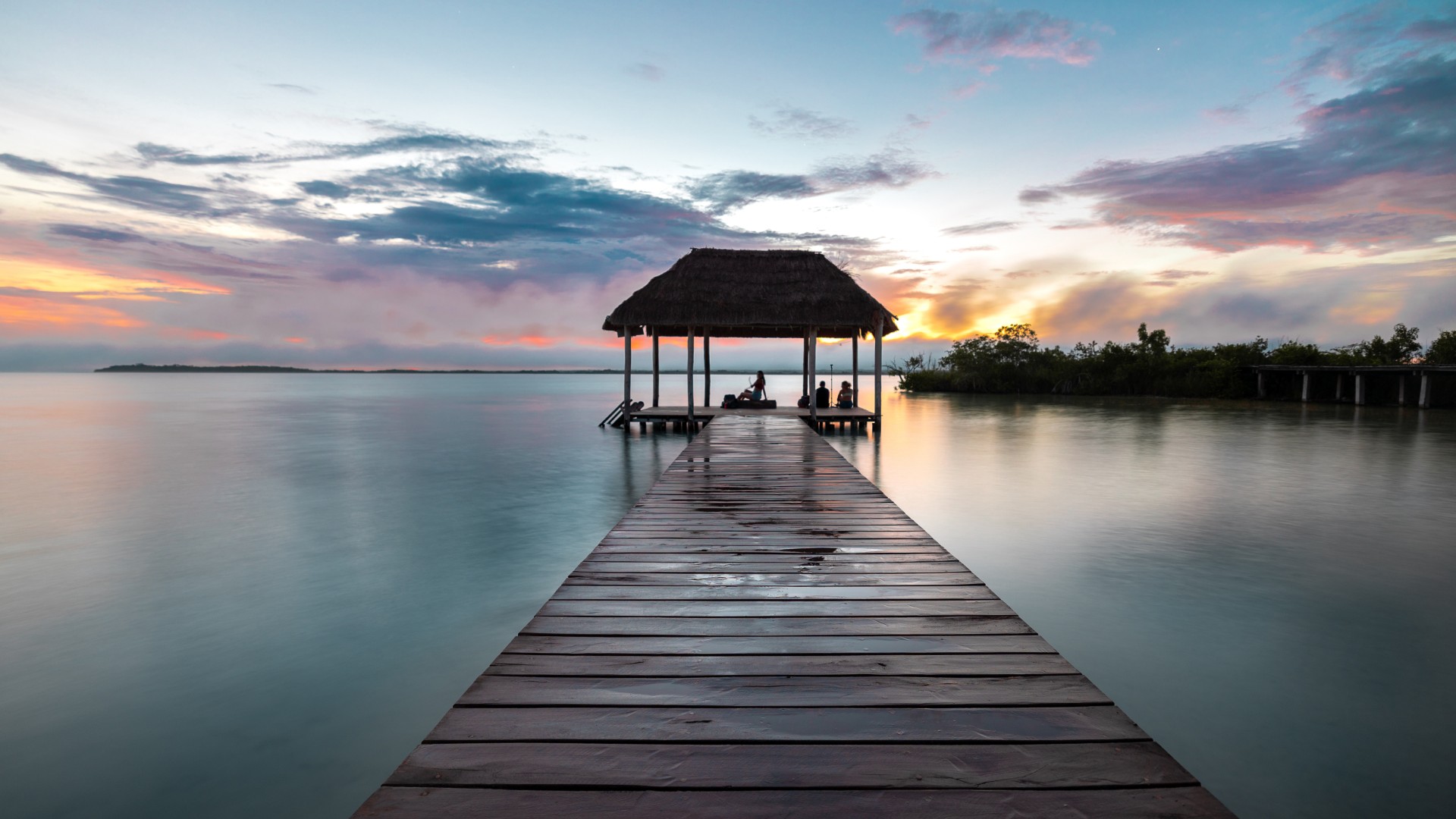 Wooden dock leading to a thatched-roof pavilion over calm water at sunset