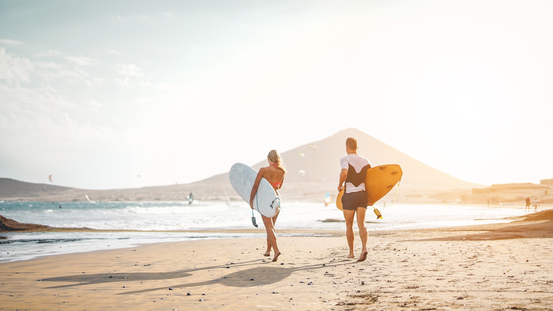 Two surfers carrying surfboards walk along a sunlit beach toward the ocean as the sun sets behind distant hills
