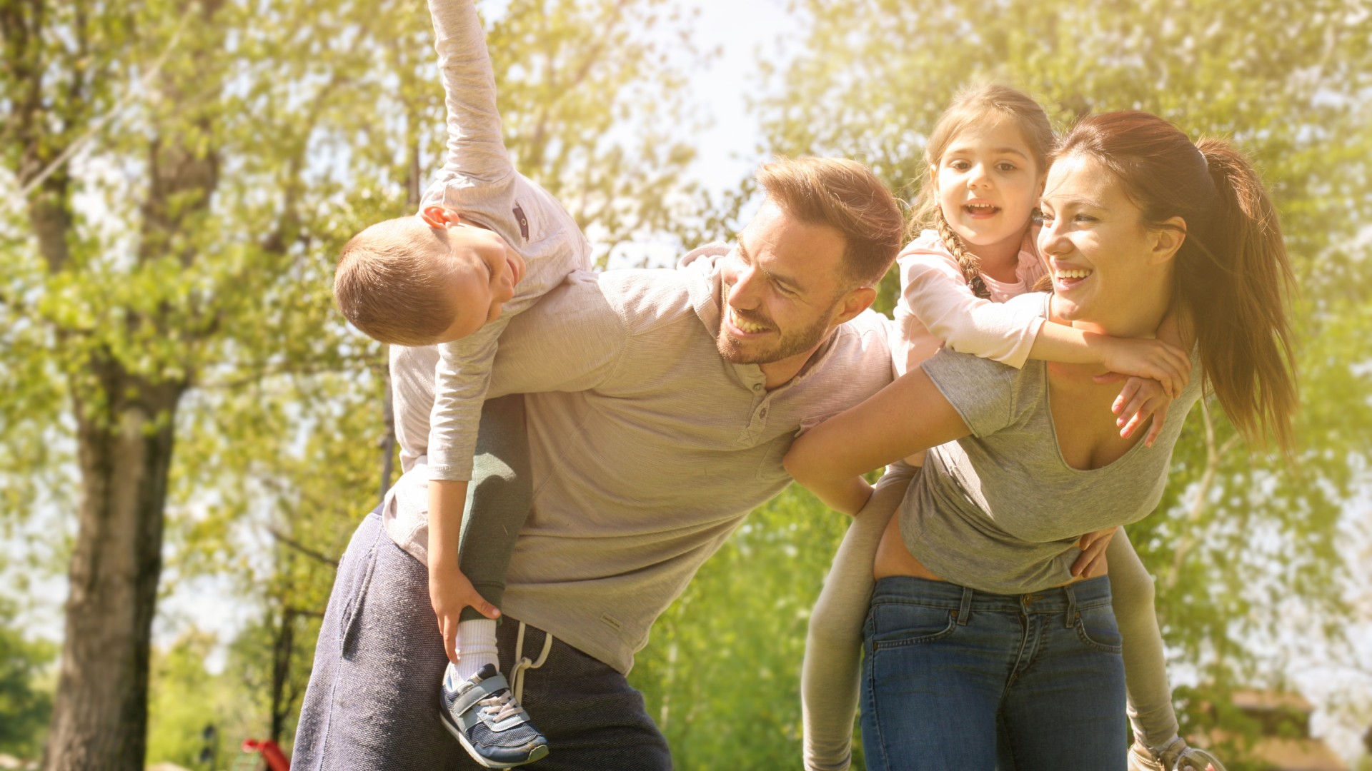 Smiling family of four playing together outdoors as parents give their children piggyback rides in a sunny park.