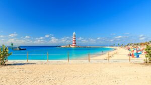 Red and white striped lighthouse overlooking a calm turquoise beach with white sand and clear blue skies.