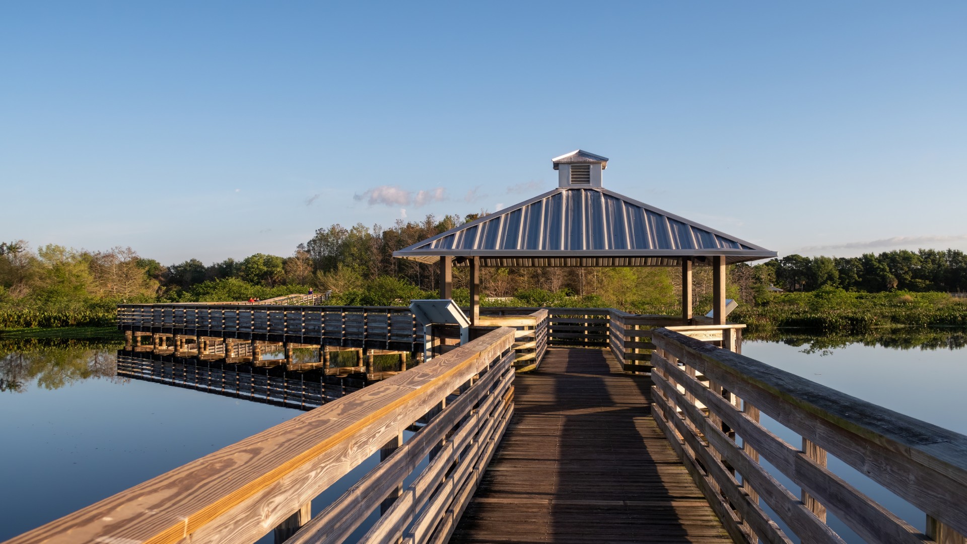 Wooden boardwalk leading to a covered observation pavilion overlooking a calm lake surrounded by lush greenery under a clear blue sky.