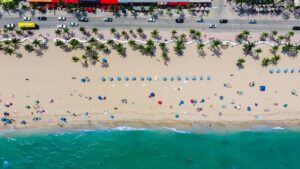 Aerial view of a sandy beach lined with palm trees, colorful umbrellas, and turquoise ocean water beside a coastal roadway.
